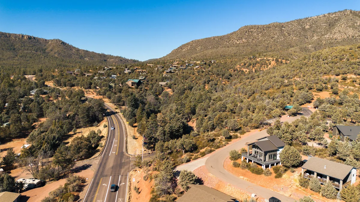 A sweeping exterior view featuring a mountainous landscape, a winding road leading through dense trees, scattered homes nestled in the greenery, and an expansive sky. The scene captures the serene, untouched beauty of the area and the call of adventure just outside the doorstep.