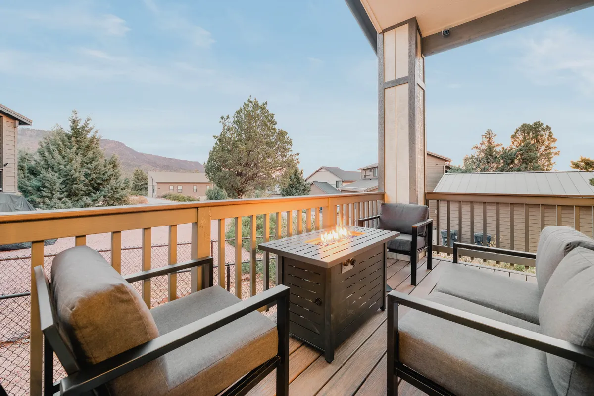 The image showcases the exterior of a modern mountain home nestled among tall pine trees. At the center, a cozy fire pit is surrounded by comfortable outdoor seating, all positioned to take in sweeping views of distant mountains. The setting sun casts a warm glow over the deck, inviting guests to relax, socialize, and savor the peaceful forest atmosphere.