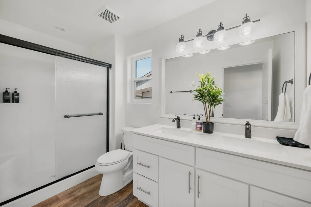 A spacious bathroom featuring a double sink vanity, large mirrored wall, glass shower area, and decorative plant accents. The space is clean, modern, and designed for relaxation.
