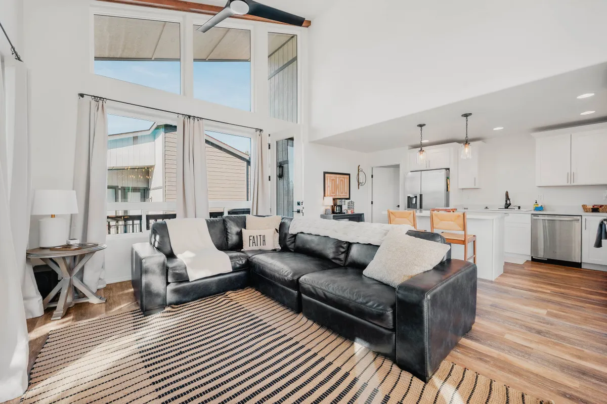 A modern living room with a black sectional sofa, large windows framing beautiful outdoor views, an open kitchen area in the background, and decorative wall art. The layout demonstrates the open-concept flow and emphasizes light-filled, inviting interiors.