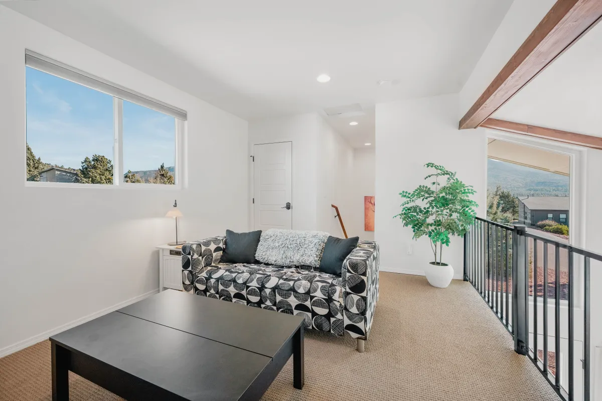 A comfortable living area featuring a black and white patterned couch and a coffee table positioned by a large window, with an indoor plant adding a touch of greenery. The window frames a view of the outdoors, hinting at the natural beauty surrounding the home.