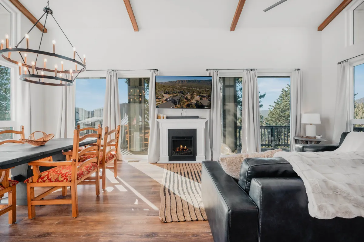 A wide view of the main living space featuring a cozy sofa, an elegant chandelier, an electric fireplace, and large windows allowing natural light to fill the room. The dining table is positioned nearby, seamlessly connecting the living and dining areas, while the overall decor strikes a perfect balance between rustic charm and modern comfort.