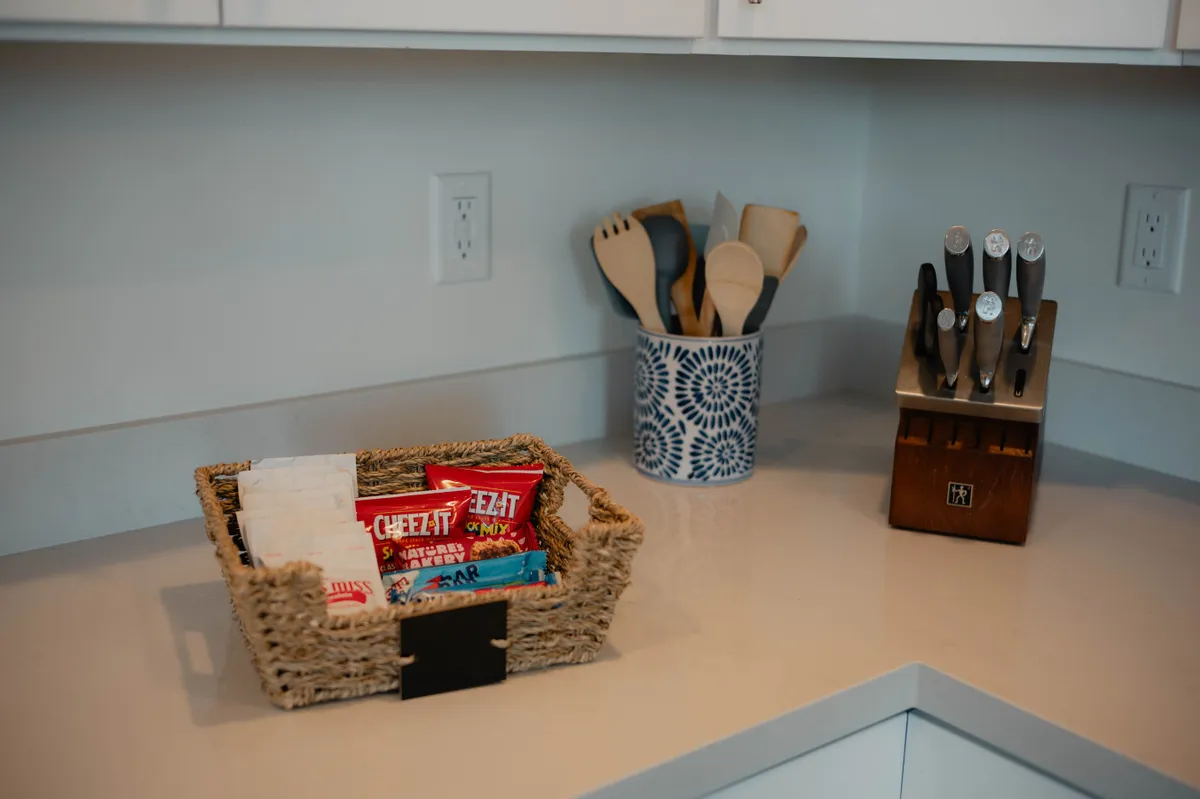 A kitchen scene with a basket of snacks, a cutting board, kitchen utensils, and a knife block is shown. The arrangement suggests an active, social kitchen environment perfect for preparing group meals or snacks together.